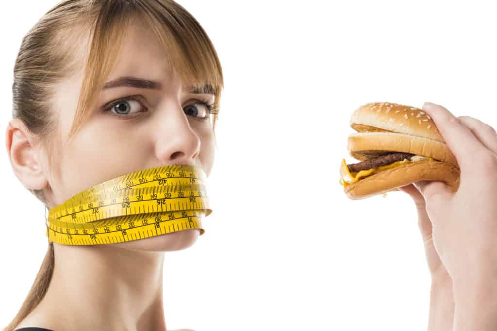 woman with measuring tape over mouth trying to eat a burger.